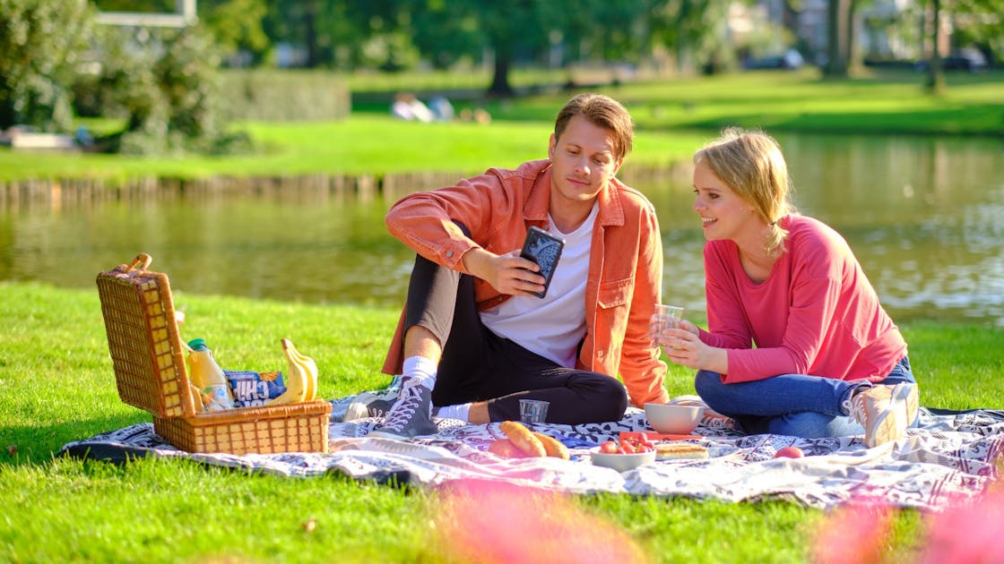 Jonge man en vrouw zijn aan het picknicken. Jongen kijkt naar zijn beleggingsrekening op zijn telefoon en de jonge vrouw kijkt mee.